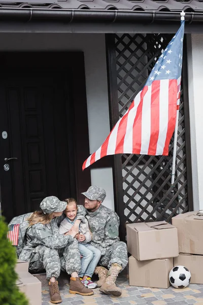 Military parents with daughter hugging, while sitting on threshold near cardboard boxes and american flag on blurred foreground — Stock Photo