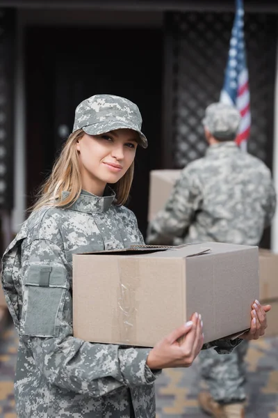 Blonde woman in camouflage holding cardboard box and looking at camera with blurred military man on background — Stock Photo
