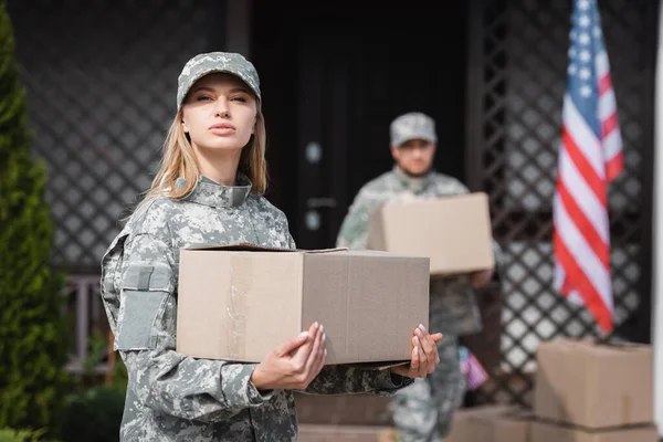 Confident woman in camouflage holding cardboard box and looking at camera with blurred military man on background — Stock Photo