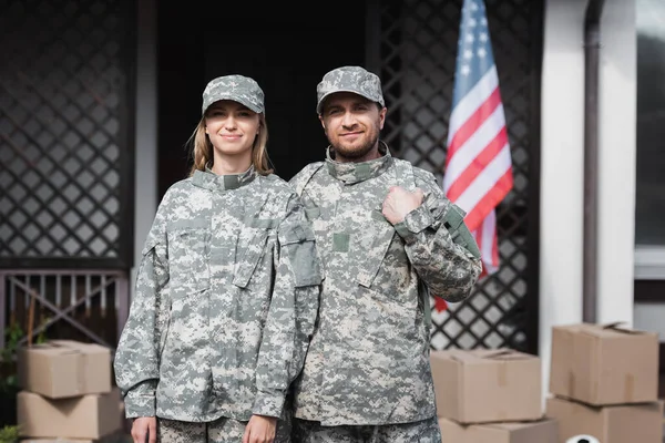 Улыбающаяся военная пара смотрит на камеру возле картонных коробок, с размытым домом на заднем плане — стоковое фото