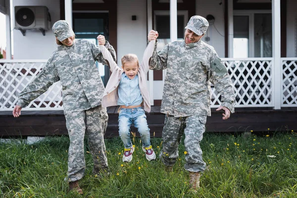Счастливые военные мать и отец поднимая дочь над травой на заднем дворе рядом с домом — стоковое фото