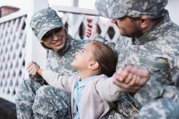 Feliz padre militar y madre tomados de la mano con su hija, mientras se sienta en el umbral de la casa en un fondo borroso - foto de stock