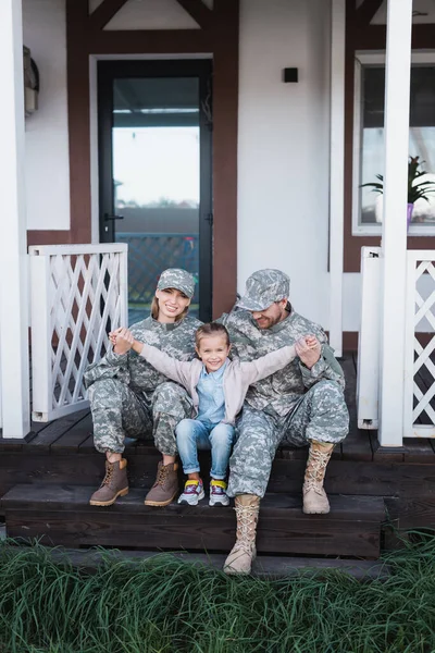 Figlia felice con le mani tese seduta vicino a madre e padre in uniformi militari sulla soglia di legno — Foto stock