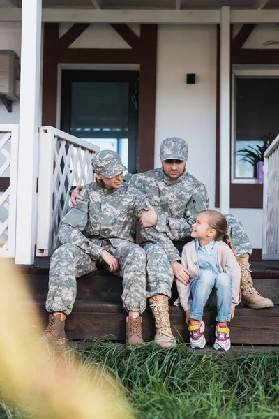 Nivel de superficie del padre militar y la madre con la hija, sentado en el umbral de la casa en primer plano borrosa - foto de stock