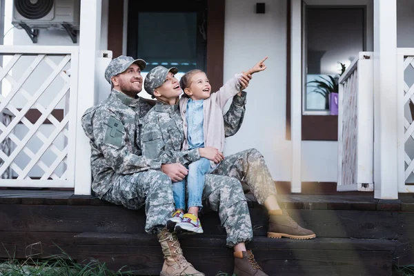 Happy military father and mother and daughter pointing with finger, while sitting on house threshold on blurred foreground — Stock Photo