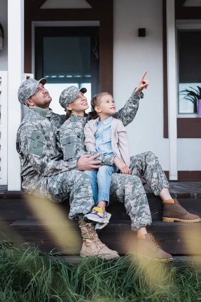 Happy military mother and father pointing with finger, looking away, sitting with daughter on house threshold on blurred foreground — Stock Photo