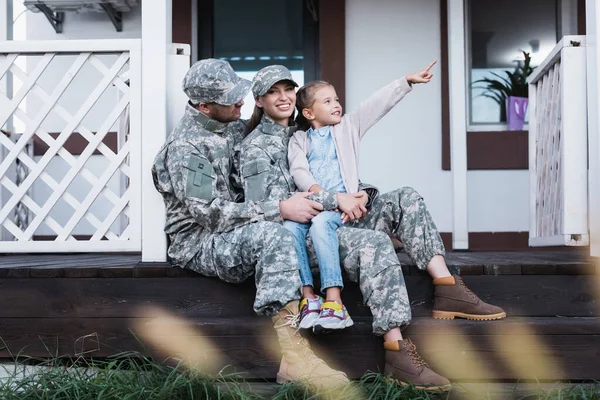 Sonriente madre militar y padre abrazándose, sentado con la hija señalando con el dedo, en el umbral de la casa en primer plano borroso - foto de stock