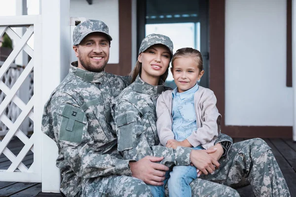 Sonriente madre militar y padre abrazándose con su hija, mientras se sienta en el umbral de la casa - foto de stock