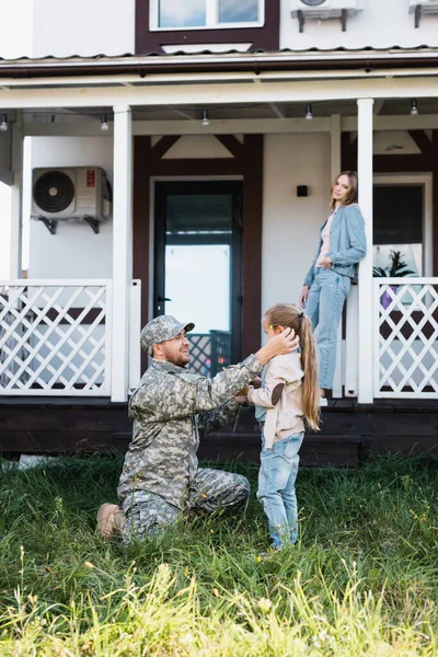 Homme militaire assis sur le genou et mettant fleur derrière fille oreille sur arrière-cour, avec femme floue et maison sur fond — Photo de stock