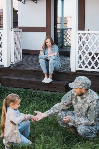 Woman with crossed arms sitting on house threshold on backyard with blurred military man and girl on foreground — Stock Photo