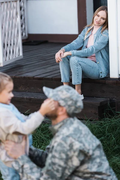 Smiling woman sitting on house threshold with blurred military serviceman and daughter on foreground — Stock Photo