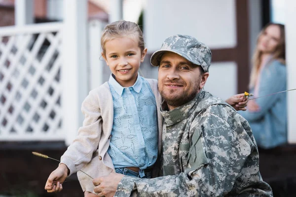 Feliz hija y padre en uniforme militar abrazando y mirando a la cámara con la mujer borrosa y la casa en el fondo — Stock Photo