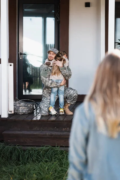 Militar sonriente abrazando a la hija, mientras está sentado en la rodilla, en el umbral con la mujer borrosa en primer plano - foto de stock
