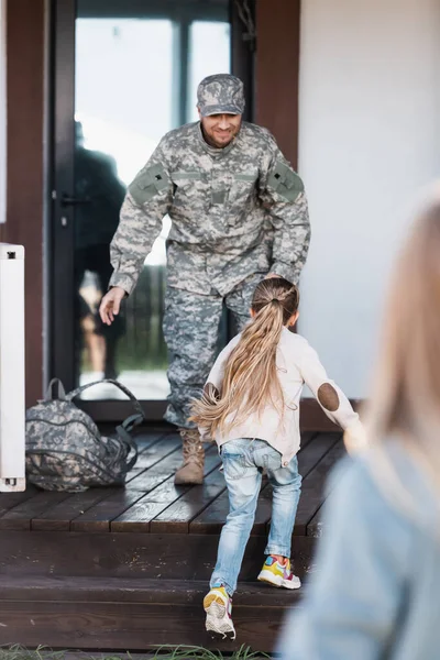 Kleines Mädchen läuft auf Militärmann zu, der auf Hausschwelle steht, mit verschwommener Frau im Vordergrund — Stockfoto