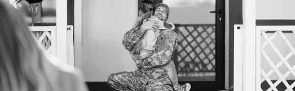 Padre in uniforme militare che abbraccia la figlia, seduto in ginocchio vicino alla porta sul retro con donna sfocata in primo piano, striscione, monocromatico — Foto stock