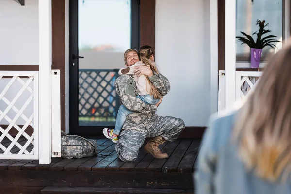 Homme de service militaire souriant embrassant sa fille, assis sur le genou sur le seuil avec une femme floue au premier plan — Photo de stock