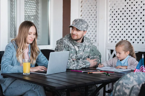 Military serviceman looking at woman typing on laptop and sitting near girl drawing with colorful pencils at home — Stock Photo