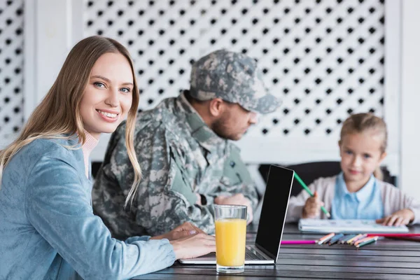 Lächelnde Ehefrau blickt in die Kamera, tippt auf Laptop, während sie am Tisch sitzt, mit verschwommenem Militärmann und Mädchen im Hintergrund — Stockfoto