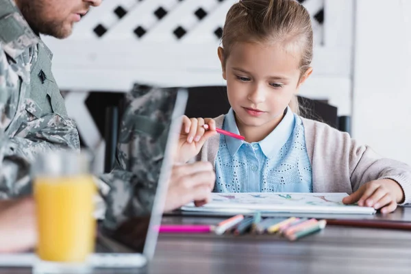 Girl with color pencil sitting near father in military uniform on blurred foreground — Stock Photo