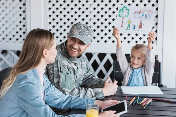 Lächelnder Mann in Militäruniform schaut Frau mit digitalem Tablet an, während er mit Tochter zu Hause sitzt und Bild zeigt — Stockfoto