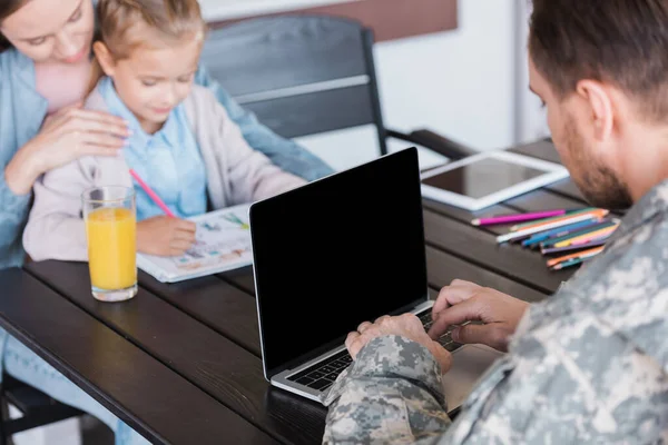 Man in military uniform typing on laptop with blank screen, while sitting at table with blurred woman and girl on background — Stock Photo