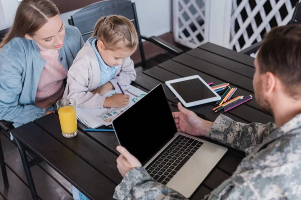Militar serviceman usando laptop e olhando para menina sentado com mulher e desenho com lápis coloridos à mesa — Fotografia de Stock
