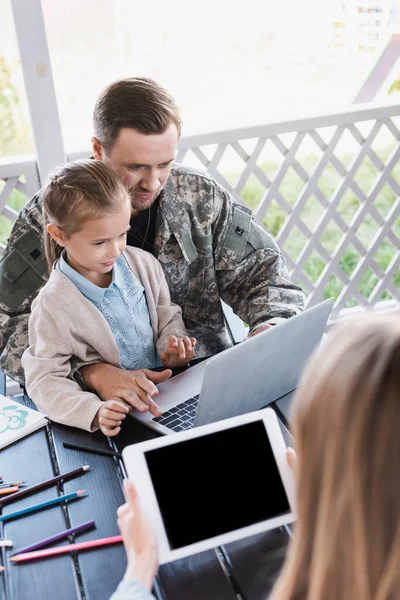 Military man hugging girl and using laptop, while sitting at table with blurred woman on foreground — Stock Photo