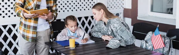 Madre atenta en uniforme militar sentada cerca de la hija escribiendo en el cuaderno, pancarta - foto de stock