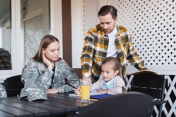 Girl writing at notebook, while sitting near father and mother at home — Stock Photo