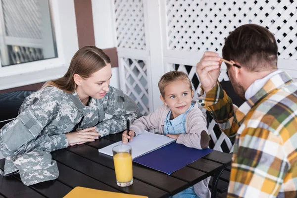 Усміхнена дівчина з блокнотом дивиться на чоловіка, який тримає олівець, сидячи біля матері у військовій формі за столом — Stock Photo
