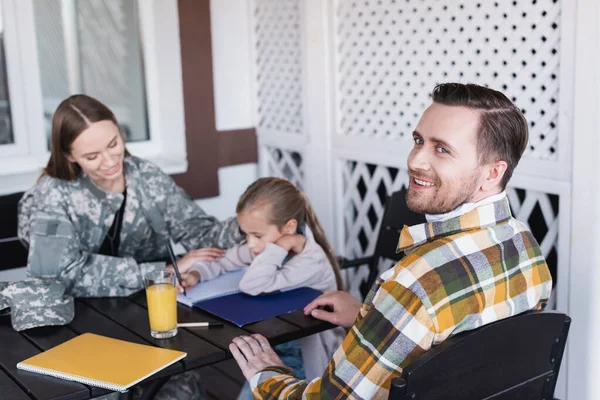 Усміхнений бородатий чоловік у картатій сорочці, сидячи за столом з розмитою жінкою та дівчиною на фоні — стокове фото