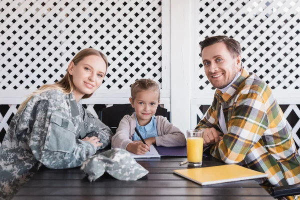 Smiling girl with man in checkered shirt and woman in military uniform sitting at table with notebooks, banner — Stock Photo