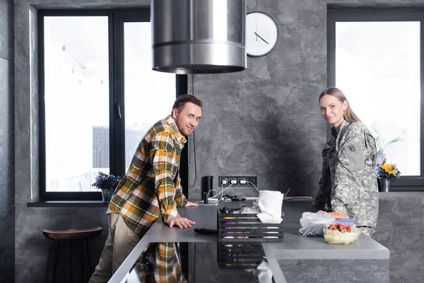 Sonriente hombre y mujer en uniforme militar mirando a la cámara, mientras se apoya en la mesa en la cocina loft - foto de stock