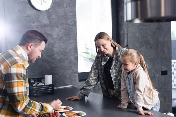 Sorridente madre e figlia appoggiate sul tavolo, mentre il padre serve piatti con frittelle in cucina — Foto stock