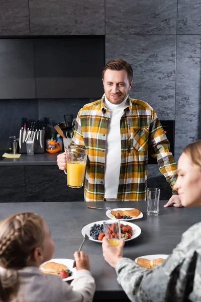 Усміхнений чоловік з глечиком стоїть біля столу з тарілками млинців та фруктами з розмитою жінкою та дівчиною на передньому плані — стокове фото