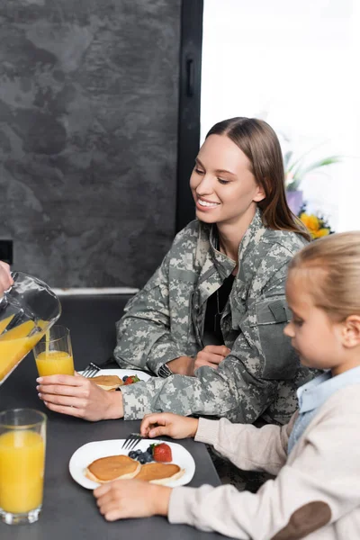 Hombre vertiendo jugo cerca de la mujer en uniforme militar y chica sentada en la mesa en la cocina - foto de stock