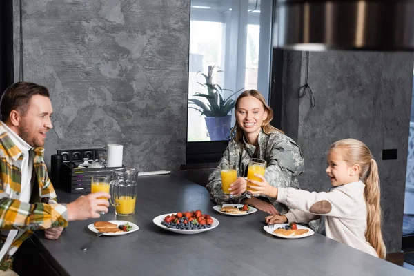 Счастливый семейный тост со стаканами сока, сидя за столом с фруктами и блинами на кухне — стоковое фото
