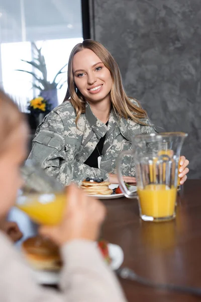 Mujer sonriente con cabello rubio sentada en la mesa con panqueques y jarra de jugo en la cocina con chica borrosa en primer plano - foto de stock