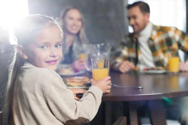 Happy girl looking at camera, while holding glass of juice and sitting at table with blurred woman and man on background — Stock Photo