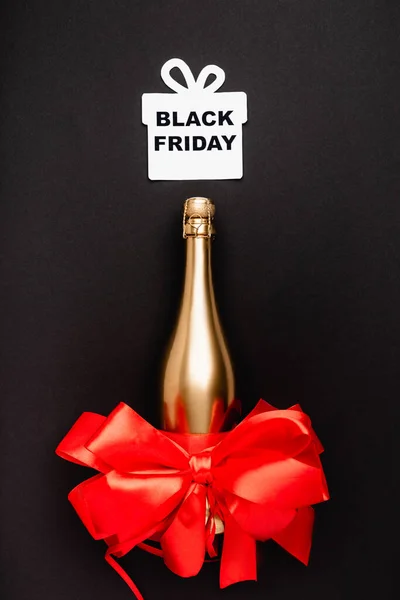 Top view of golden bottle of champagne, red bow and paper gift with black friday lettering on black background — Stock Photo