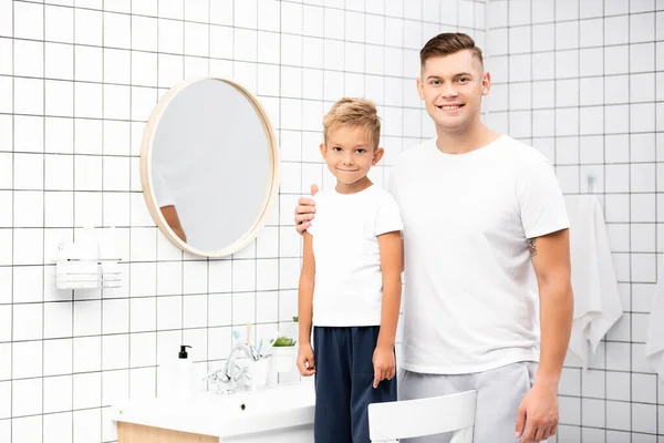 Sonriente padre abrazando hijo de pie en la silla cerca del espejo y fregadero en el baño - foto de stock