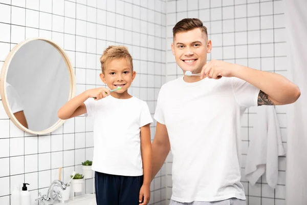 Alegre padre e hijo con cepillos de dientes mirando a la cámara en el baño - foto de stock
