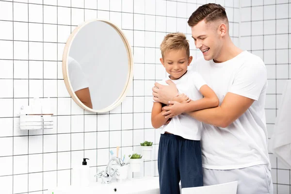 Щасливий батько обіймає сина, що стоїть на стільці біля раковини і кругле дзеркало у ванній — стокове фото