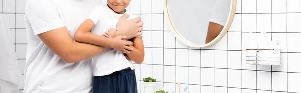 Vista recortada del padre abrazando sonriente hijo cerca de espejo redondo en el baño, pancarta - foto de stock