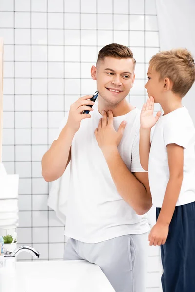 Усміхнений батько з електричною бритвою, що стоїть біля сина, торкається щоки у ванній кімнаті — стокове фото