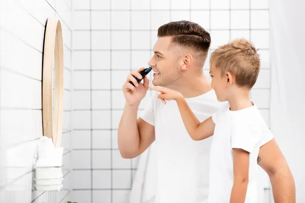 Hijo señalando con el dedo mientras mira al espejo, de pie cerca de padre sonriente con maquinilla de afeitar eléctrica en el baño - foto de stock