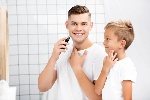 Alegre padre con maquinilla de afeitar eléctrica mirando a la cámara mientras está cerca de su hijo mirando el espejo en el baño - foto de stock