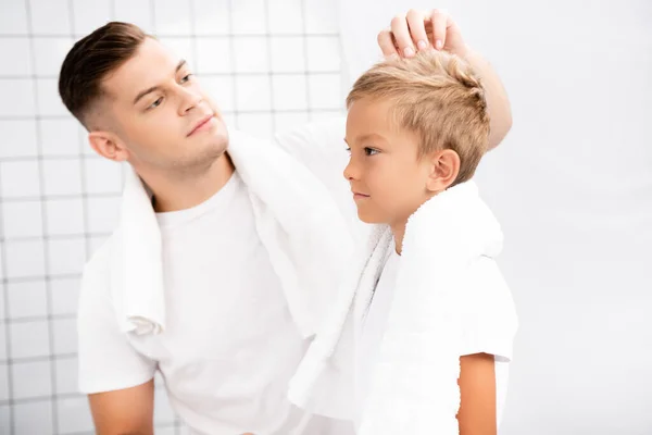 Padre con asciugamano sopra il collo in piedi vicino e fissare i capelli del figlio in bagno su sfondo sfocato — Foto stock