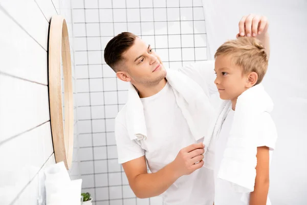 Vater mit Handtuch über dem Hals fixiert Haare des Sohnes beim Blick auf Spiegel im Badezimmer auf verschwommenem Hintergrund — Stockfoto