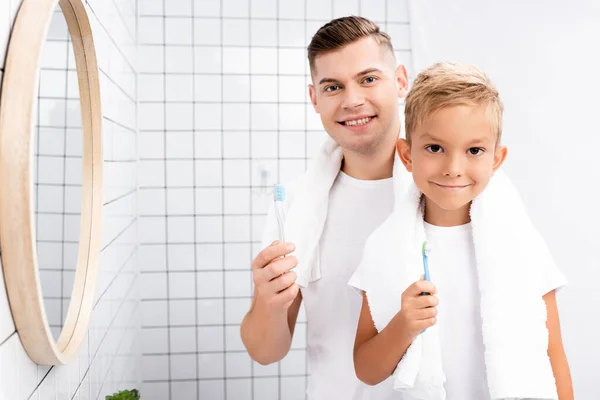Sonriente padre e hijo con cepillos de dientes mirando a la cámara cerca del espejo en el baño - foto de stock